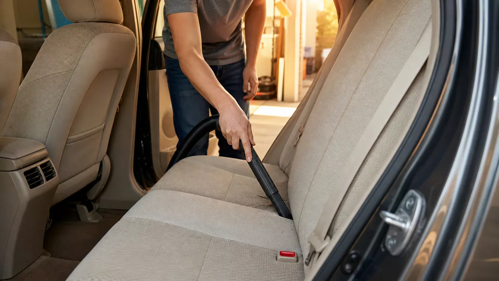 Crevice vacuum tool removing dark pet hair from the seam of a beige cloth car seat in a home garage
