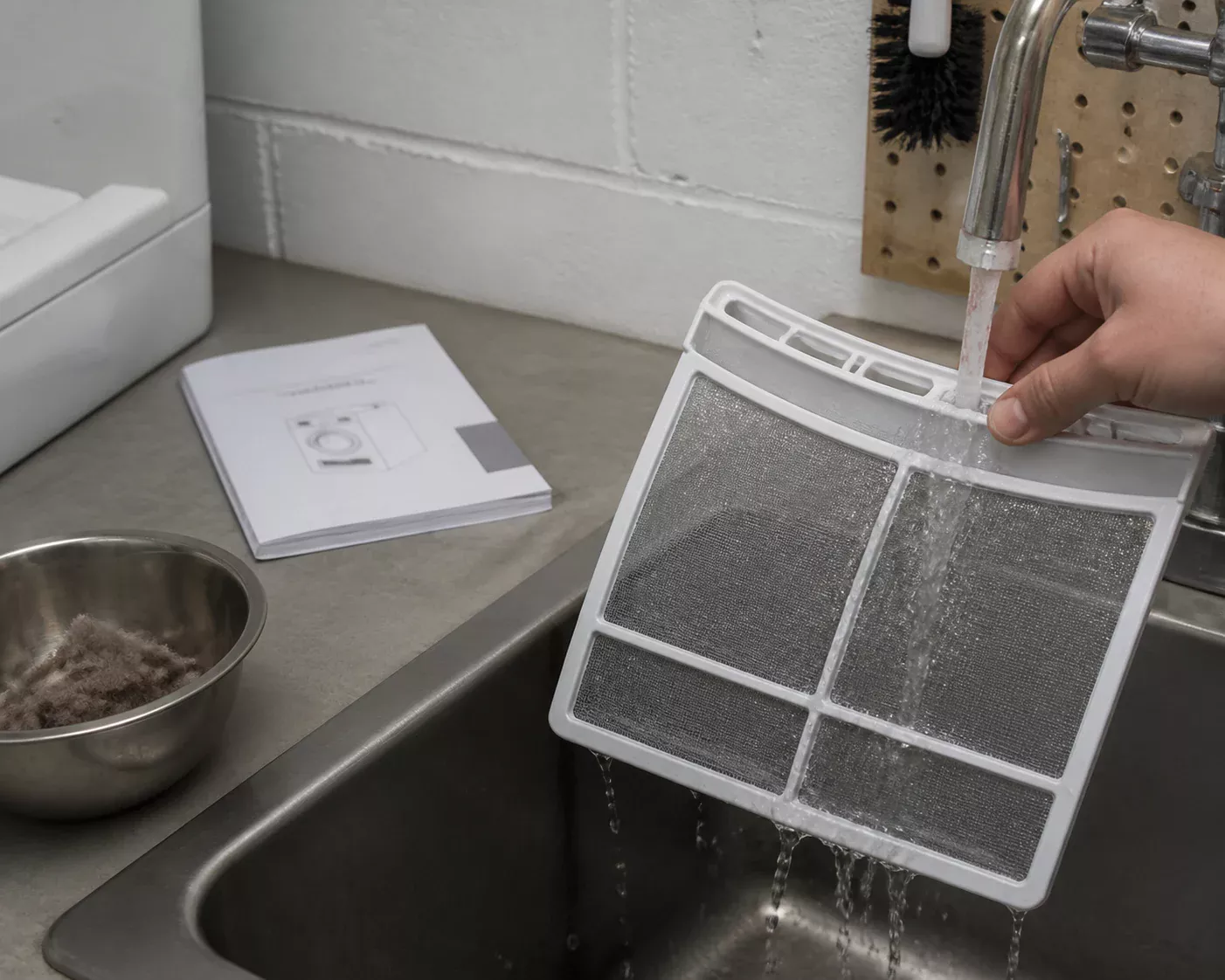 A removable appliance filter being rinsed at a stainless utility sink beside a closed washing machine manual