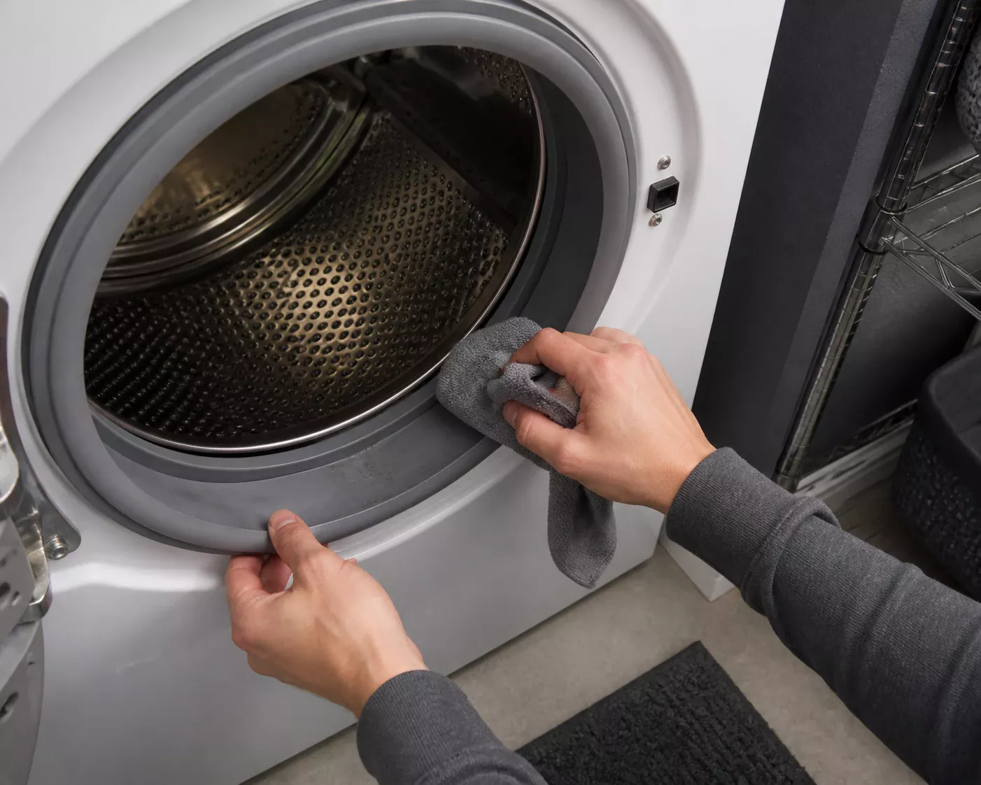 Hands wiping the rubber gasket and stainless drum of an empty front-loading washing machine with a gray microfiber cloth